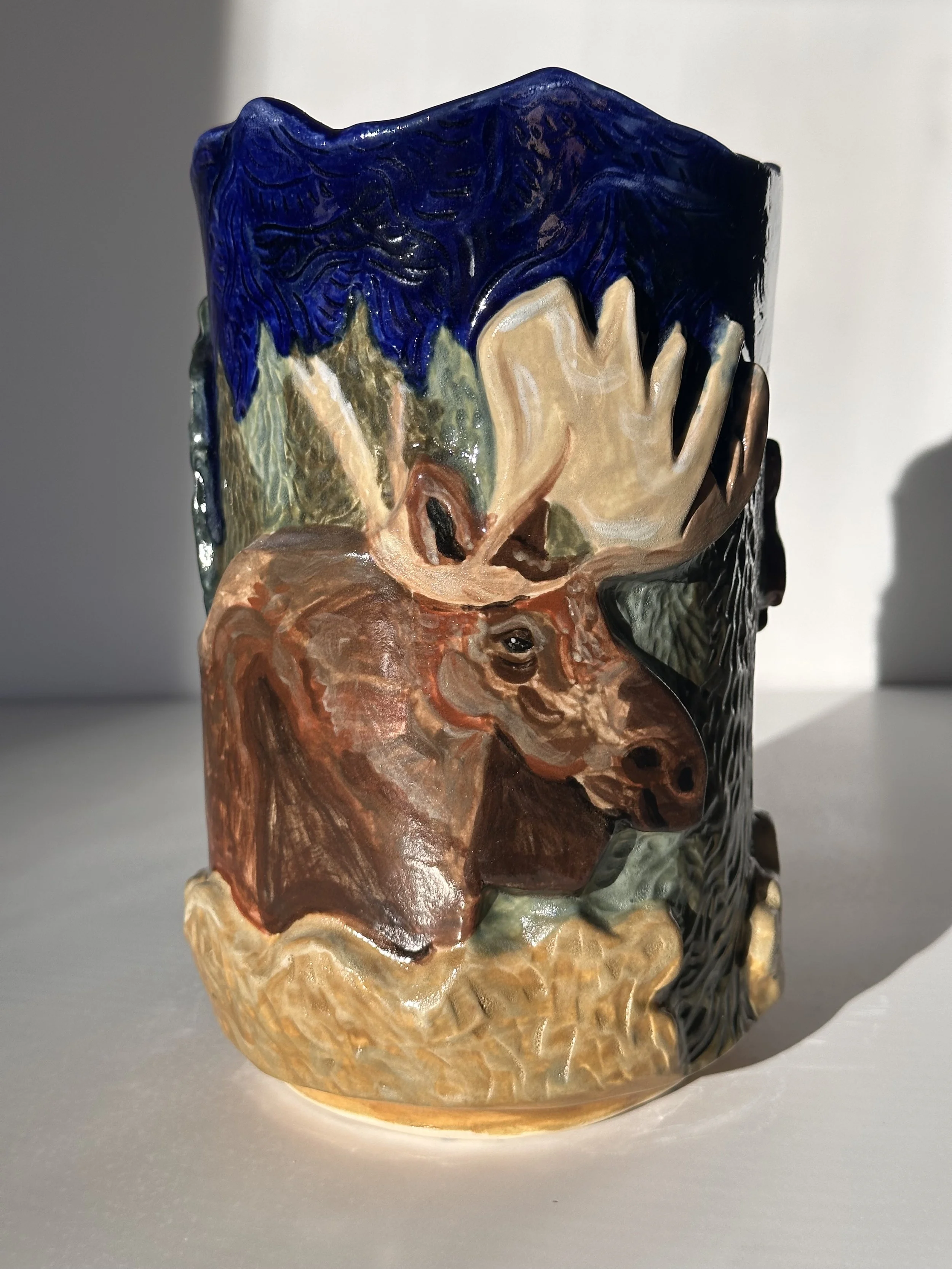 A ceramic vase with a colorful, painting of a moose with large antlers, painted in earthy tones, with blue, black, and beige accents.