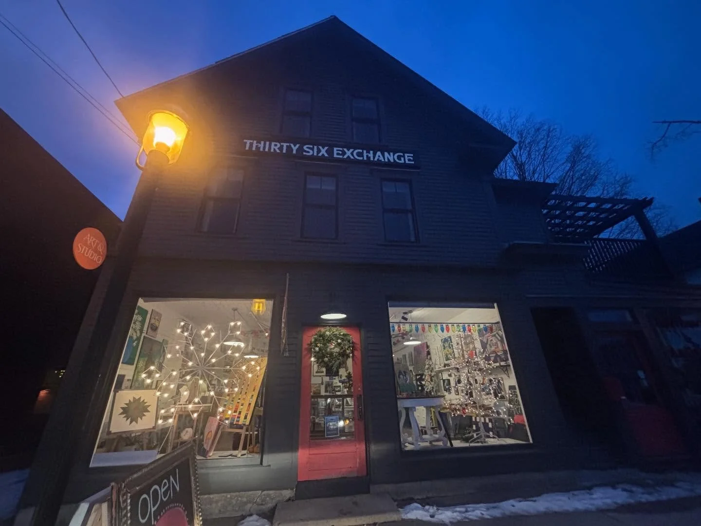 A three-story building with a sign that reads 'Thirty Six Exchange.' The ground floor has a shop with large windows displaying various items and decorations, lit from inside. There is a lit street lamp on the left side, and a small sign outside indicates the store is open. Snow is on the ground, suggesting wintertime.