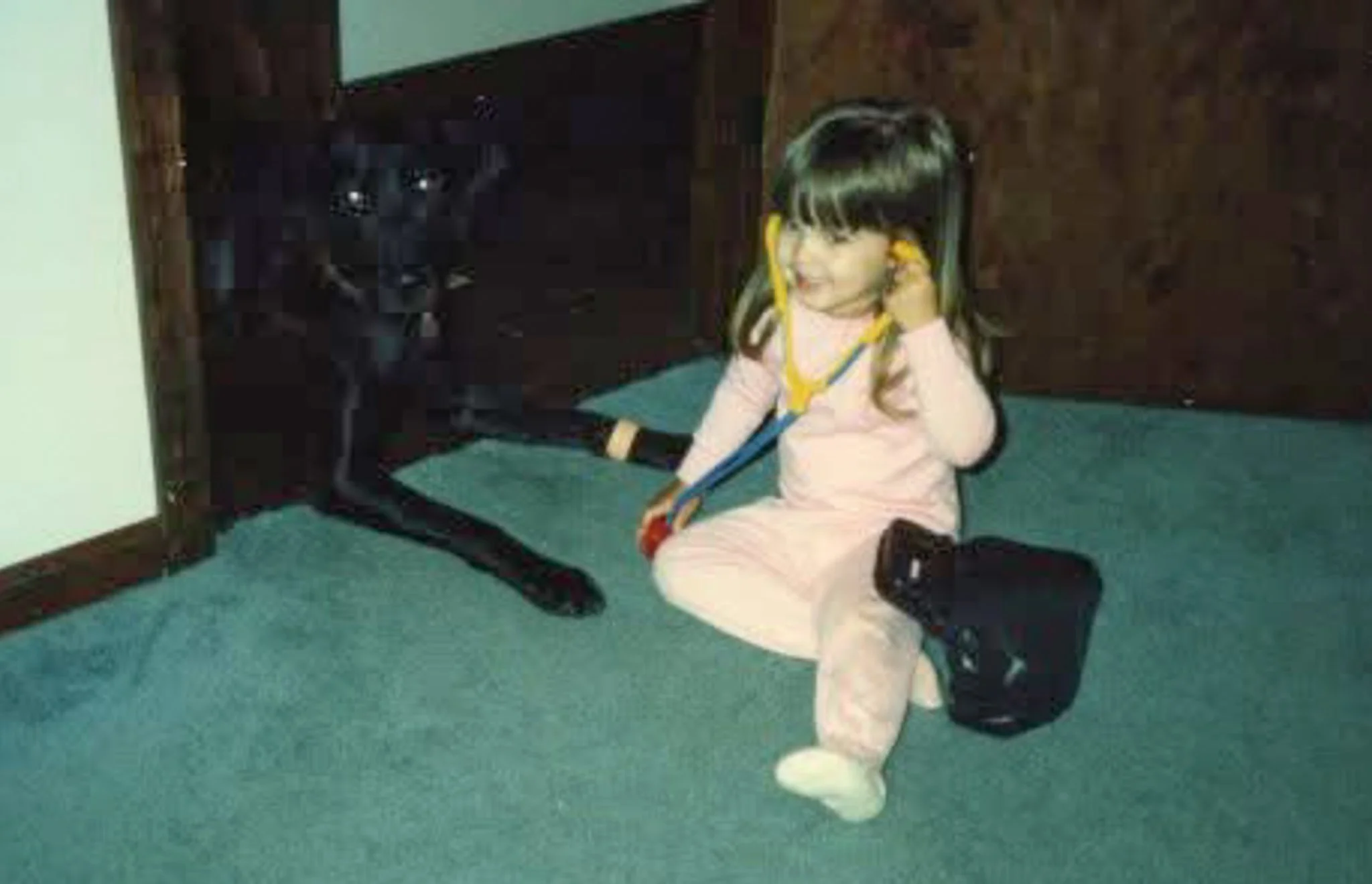 A young girl sitting on a teal carpeted floor, holding a toy medical kit next to a black Labrador retriever. The girl is wearing pink pajamas and has long hair with bangs.
