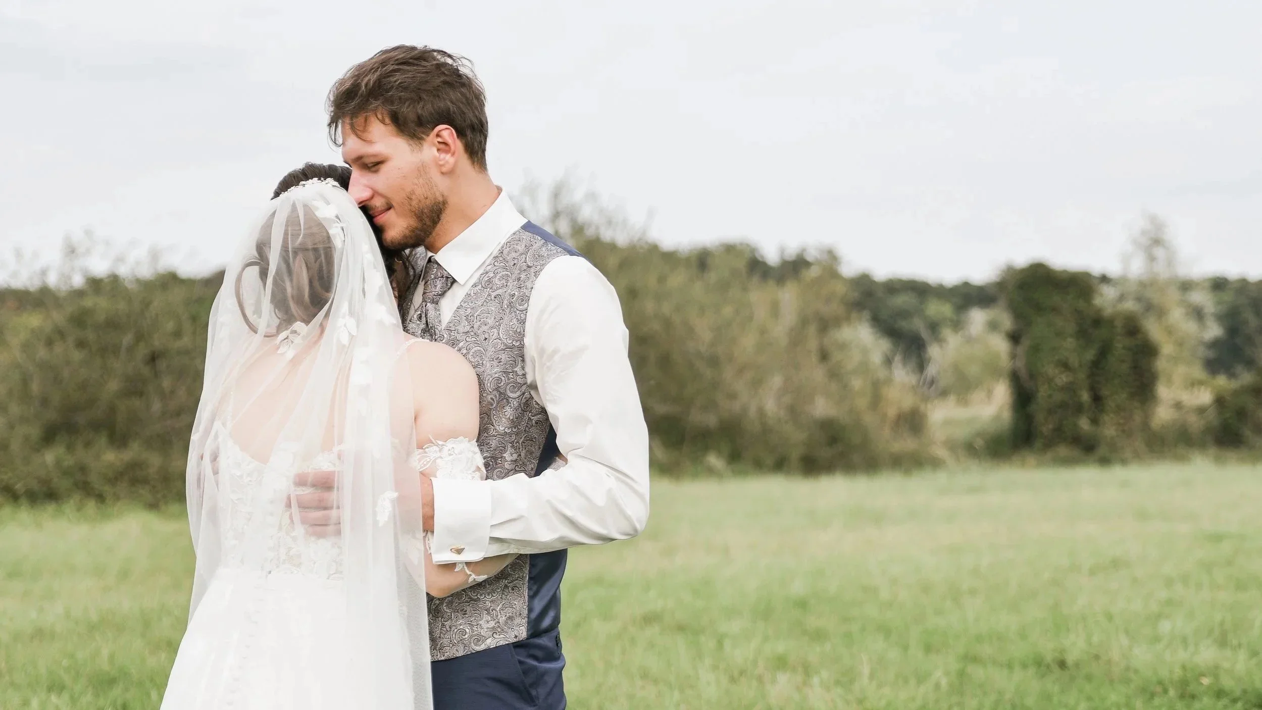Ein Brautpaar umarmt sich im Freien bei einer Hochzeit, das Paar trägt formelle Kleidung, die Umgebung ist grün und ländlich, mit Bäumen im Hintergrund.