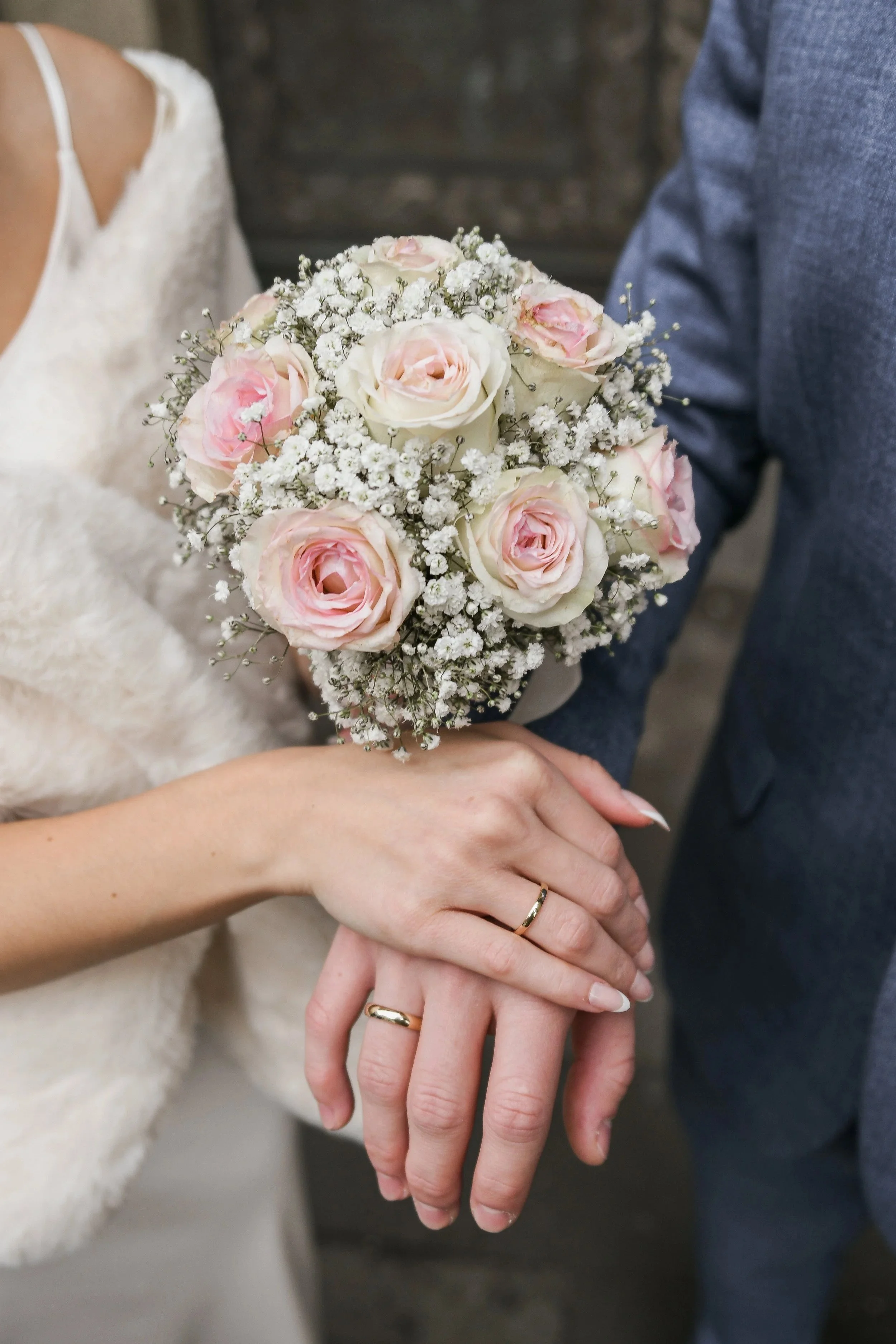 Ein Brautpaar zeigt ihre Eheringe und hält eine Hochzeitsblume mit rosa und weißen Rosen sowie kleinen weißen Blüten.