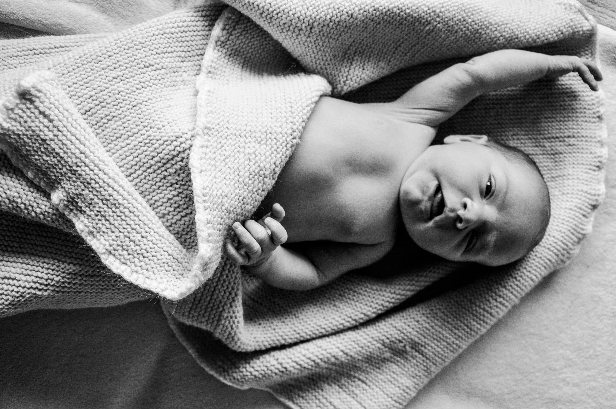 Ein neugeborener Junge liegt auf einem gefalteten Handtuch, halb bedeckt von einem Wolltuch, und schaut fragend in die Kamera.