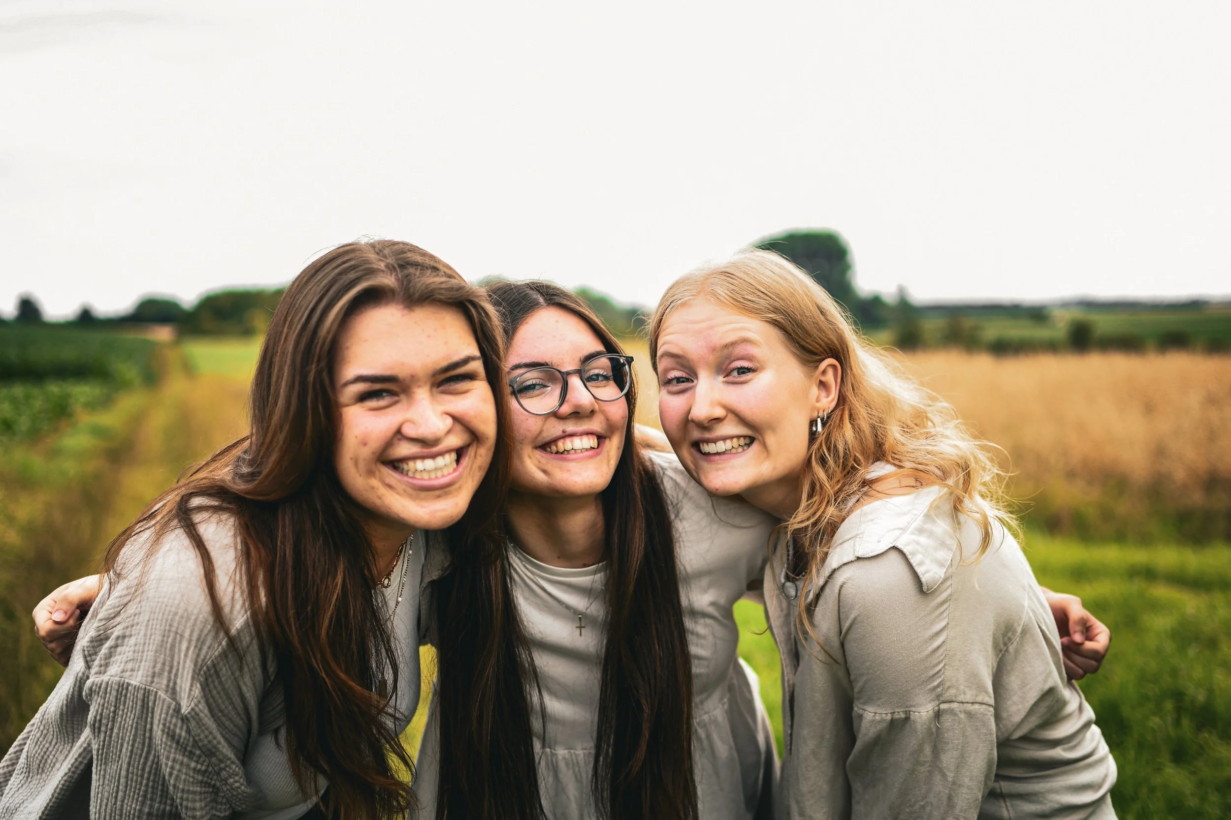 Drei lachende Mädchen stehen im Freien auf einem Feld mit grünen und goldbraunen Pflanzen im Hintergrund.