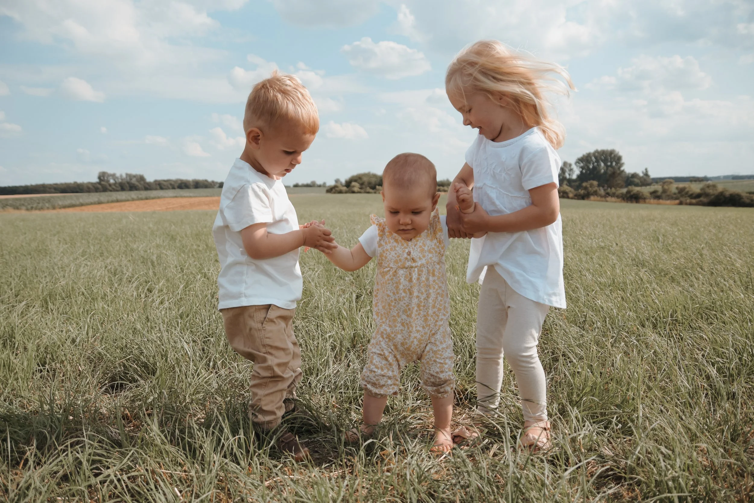 Drei kleine Kinder, ein Junge und zwei Mädchen, spielen zusammen in einem grünen Feld unter einem blauen Himmel mit Wolken.