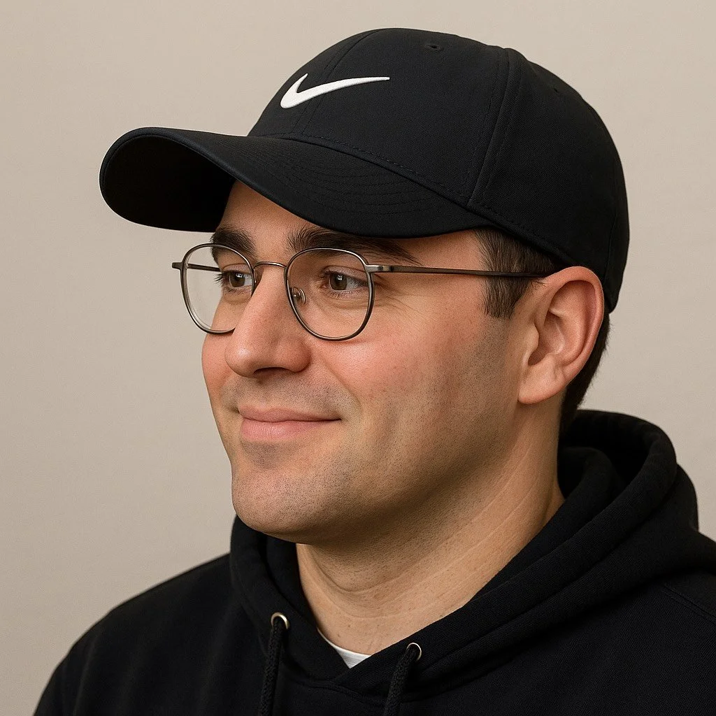 Close-up of a man wearing a black Nike cap, round glasses, and a black hoodie, smiling softly.