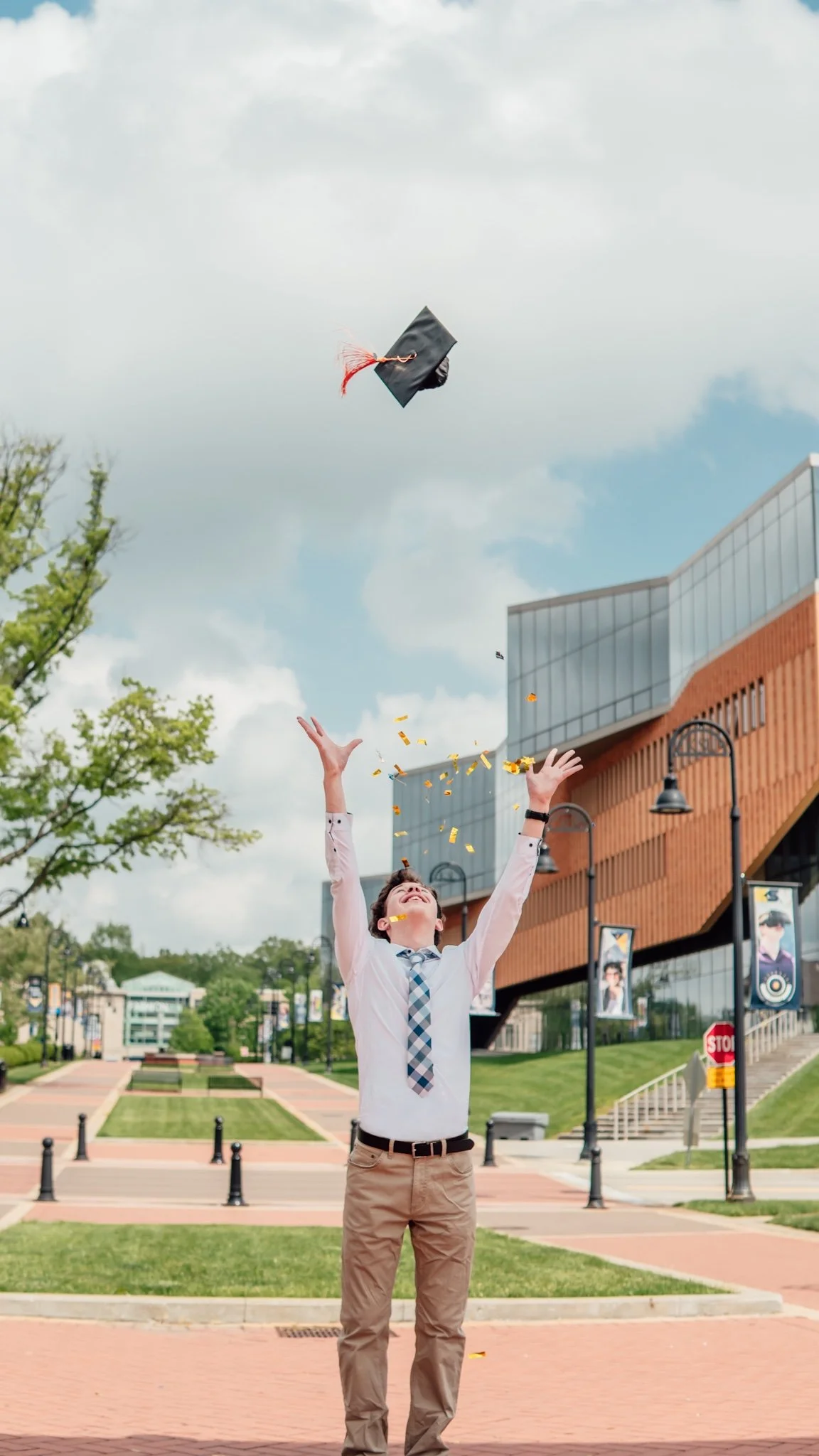 A young man in business casual attire celebrates as he throws his graduation cap into the air, surrounded by falling gold confetti, standing outdoors in front of a modern building on a college campus.