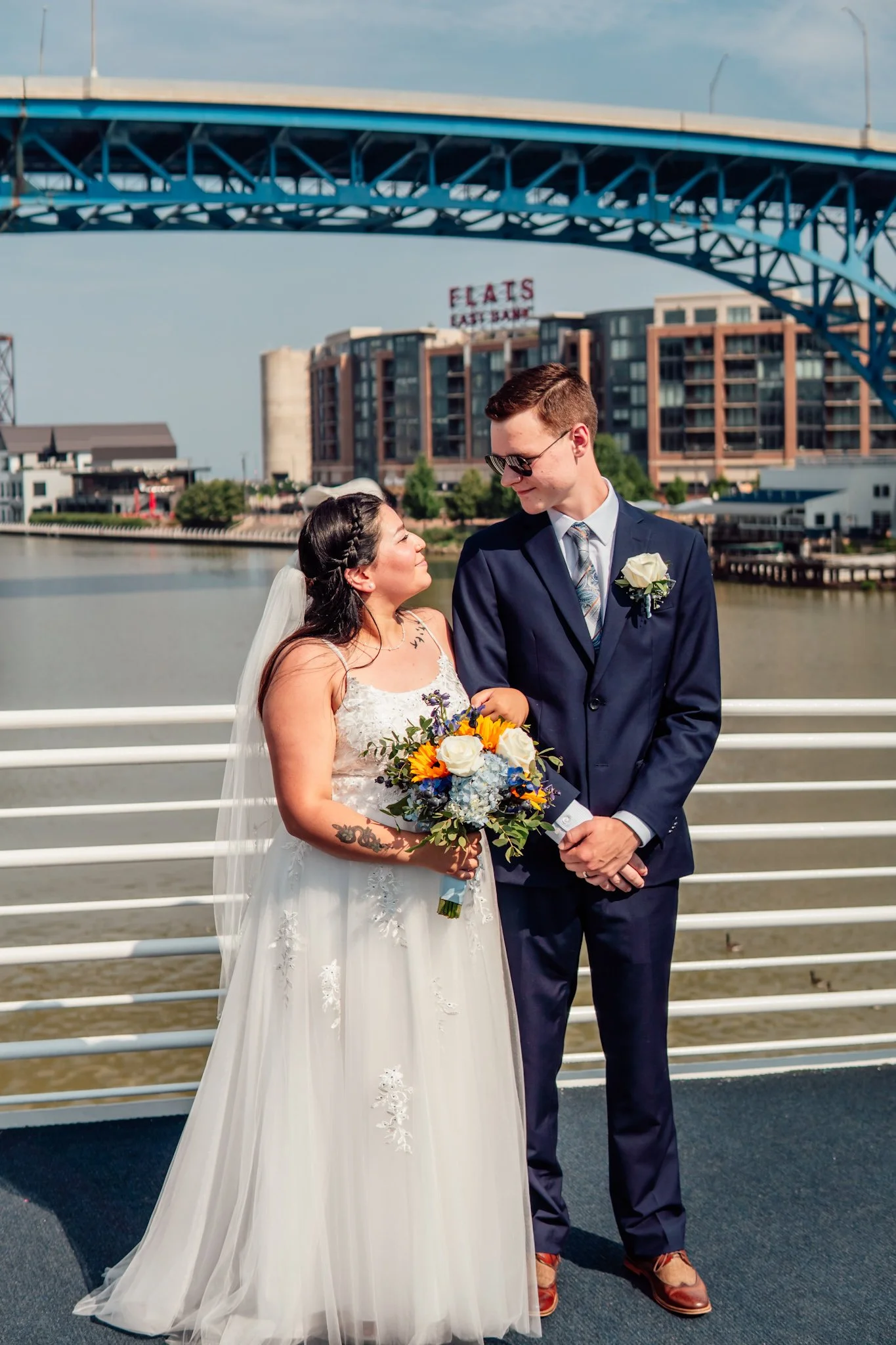 A bride and groom on a waterfront with a bridge overhead and city buildings in the background, during a wedding photoshoot.