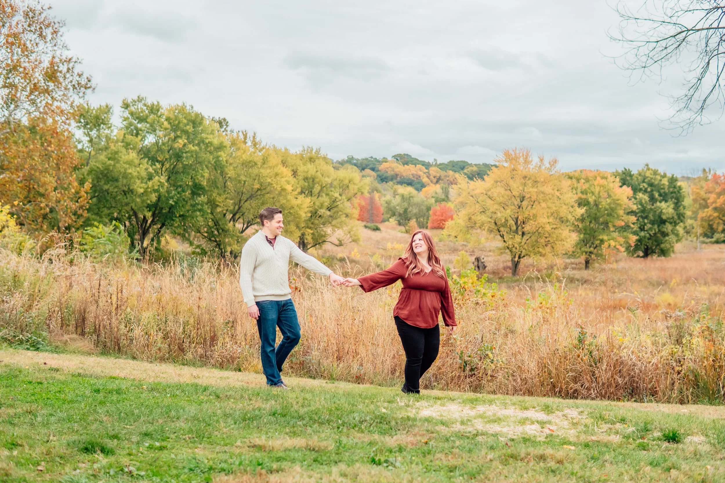 A couple holding hands and walking in a park with fall foliage on trees and a grassy path.