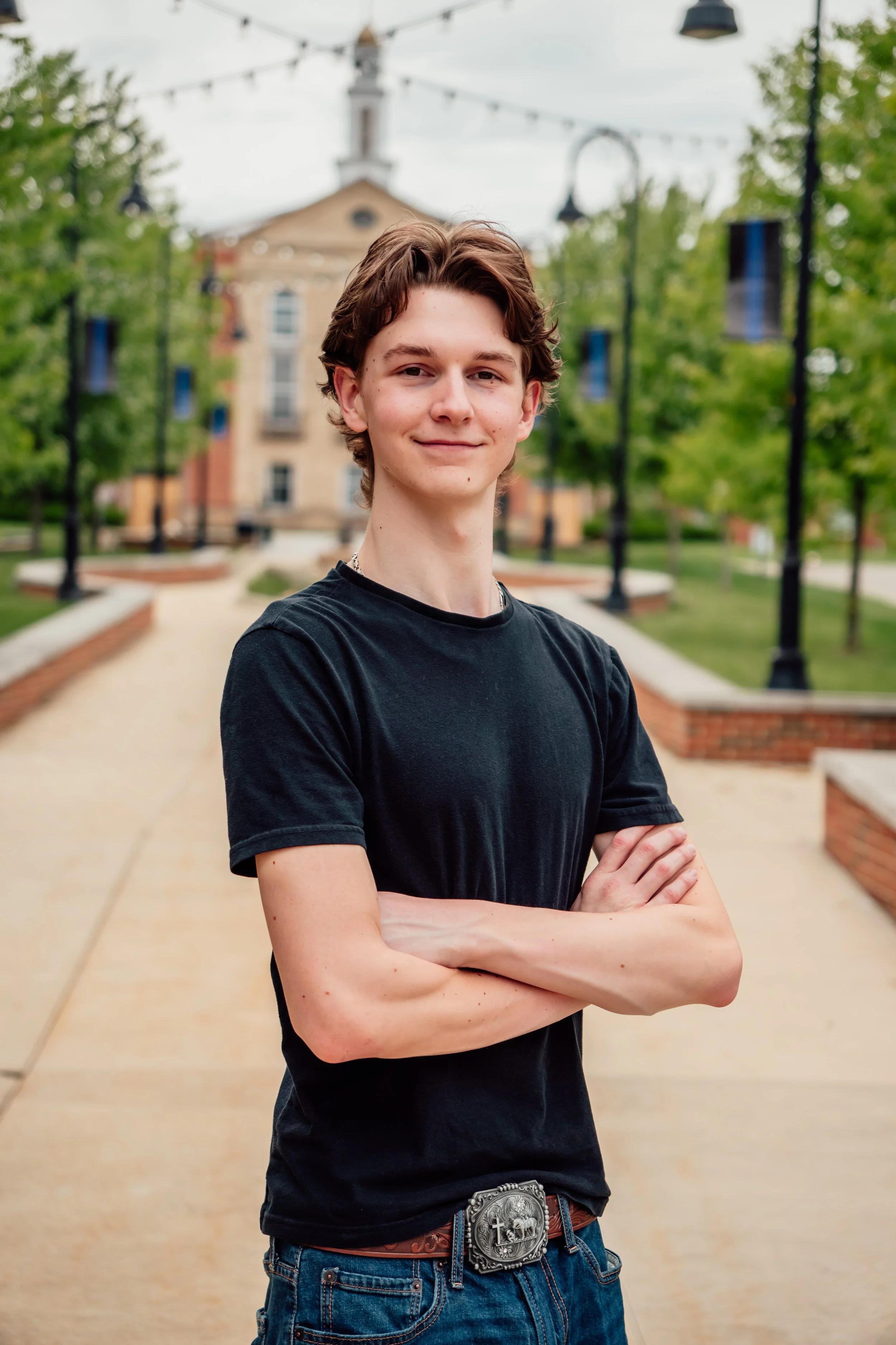 A young man with brown hair, wearing a black t-shirt and jeans, stands outdoors with arms crossed, smiling at the camera. Behind him is a brick path, green trees, and a building with a clock tower. String lights are hanging overhead.