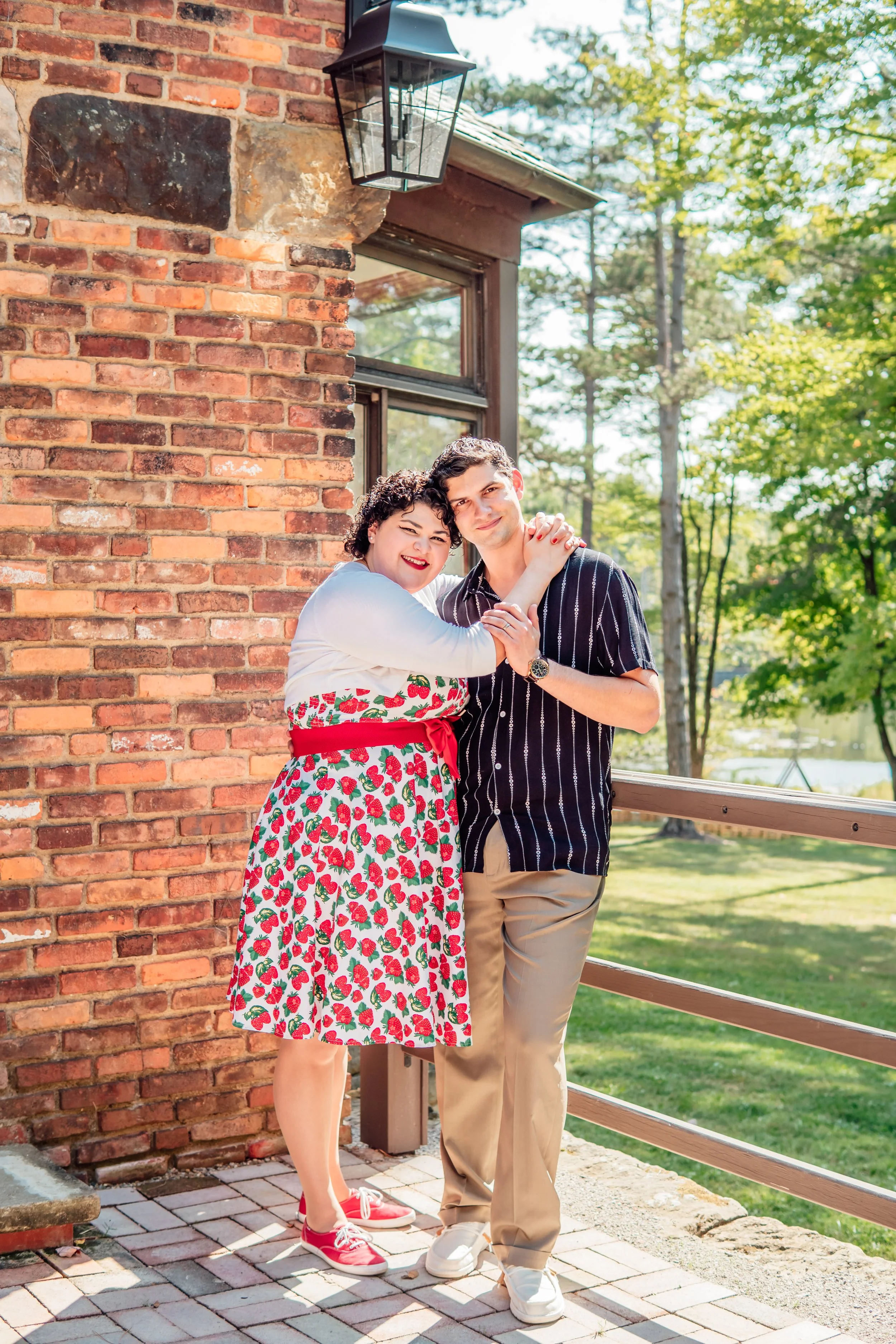 A young couple stands close together on a patio with a brick wall and a window, smiling and embracing outdoors on a sunny day with trees in the background.