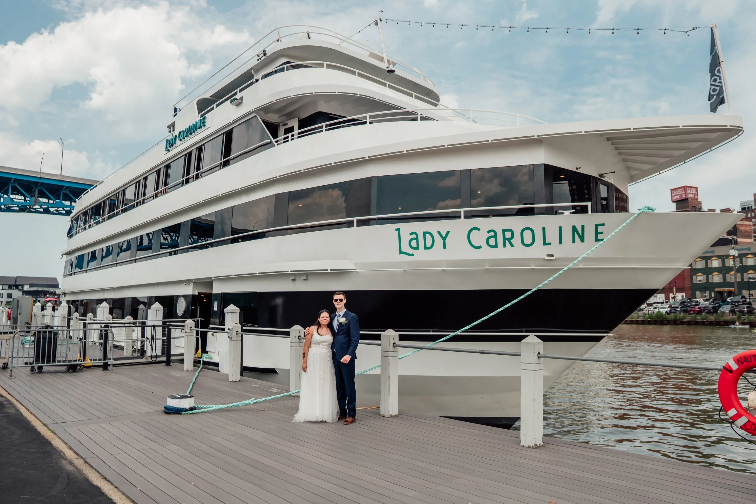 A newlywed couple standing on a dock next to a large white boat named Lady Caroline, with water and buildings in the background.