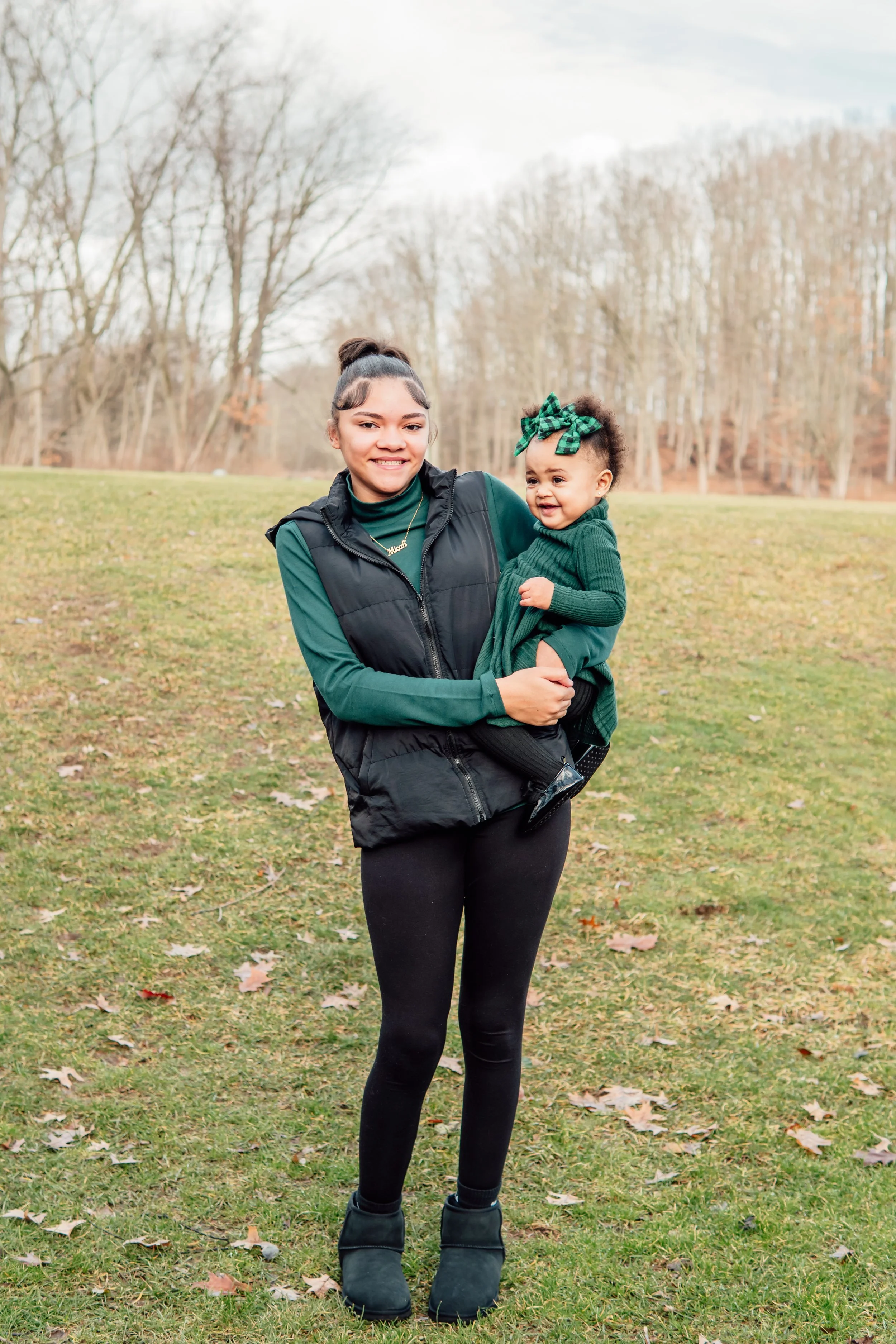 A woman holding a smiling toddler outdoors in a park during fall, with leafless trees and grass in the background.
