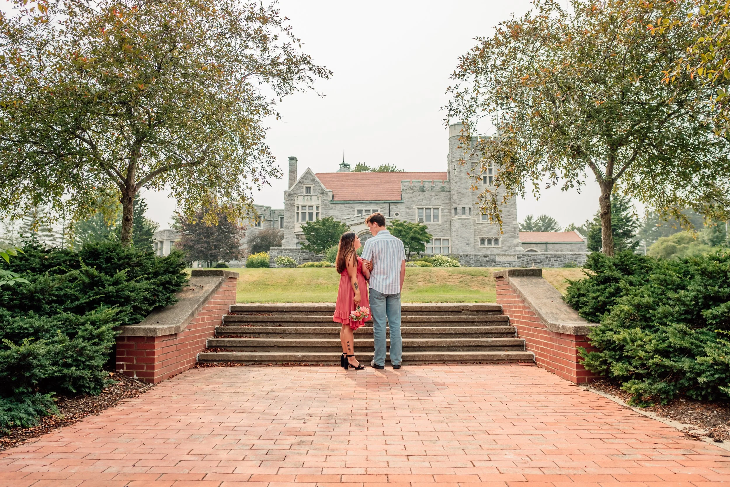 A couple dressed in semi-formal attire stands on brick steps holding hands on a cloudy day, in front of a large old mansion surrounded by trees and greenery.