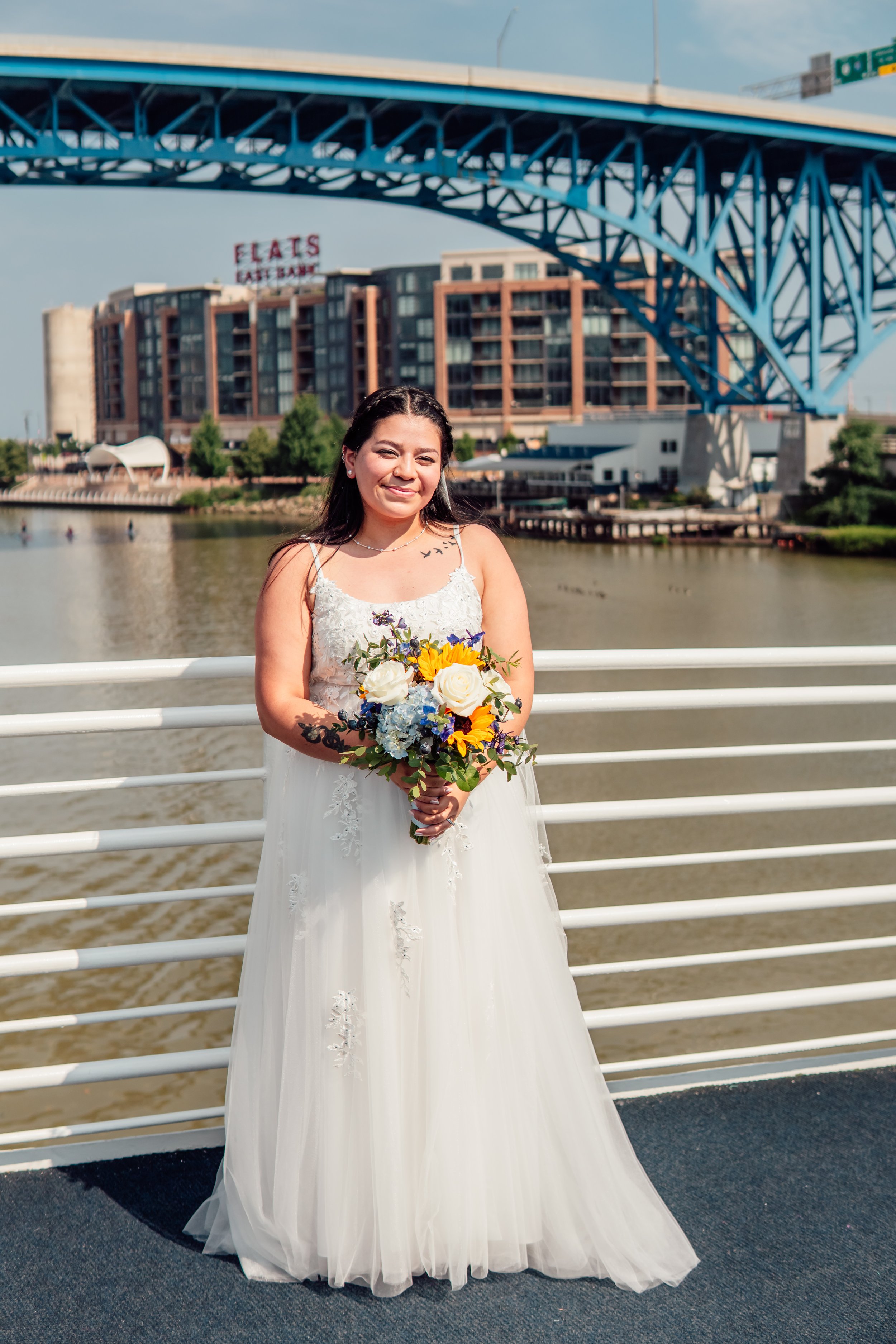 A woman in a white wedding dress holding a bouquet of flowers, standing on a riverfront promenade with a large blue bridge and city buildings in the background.
