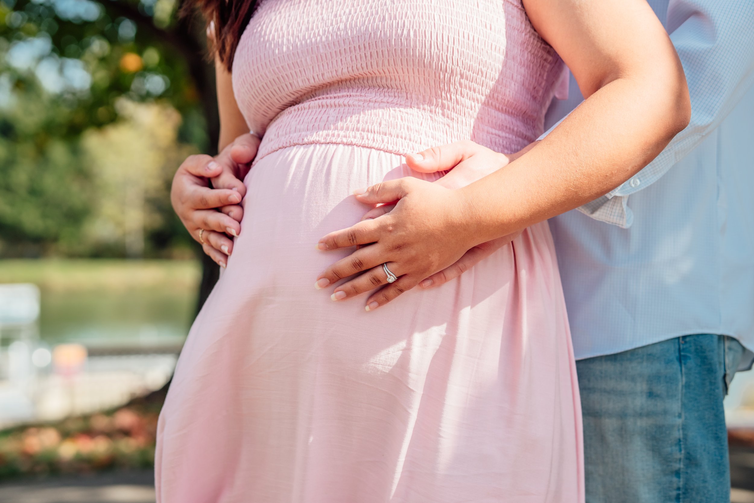A pregnant woman wearing a pink dress and a man in a light blue shirt in an outdoor setting. The woman is cradling her baby bump with both hands, one of which has a wedding ring. The background shows trees and a body of water.