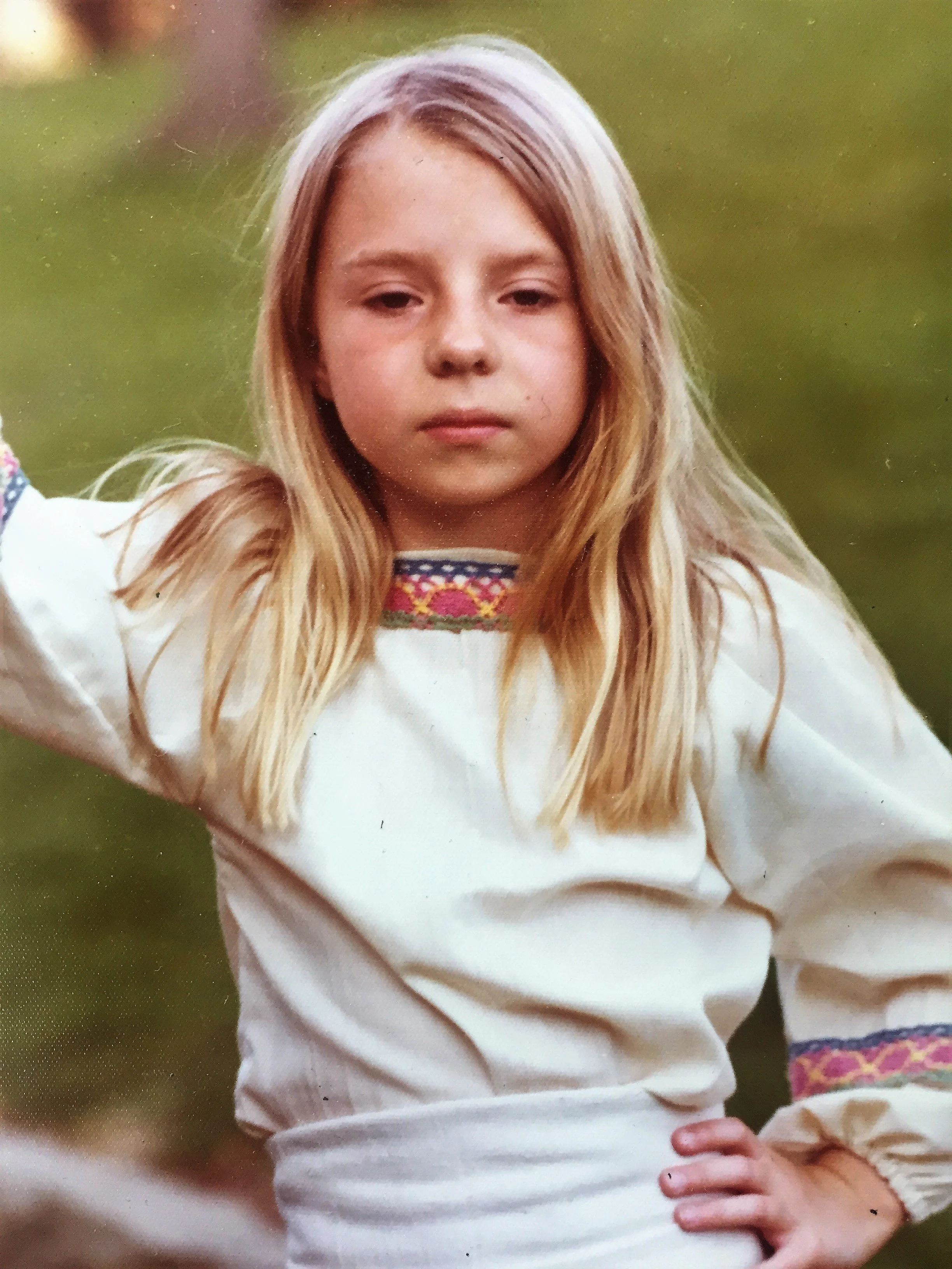 Blonde-haired girl with a enigmatic expression wearing an ecru top with colorful crocheted trim