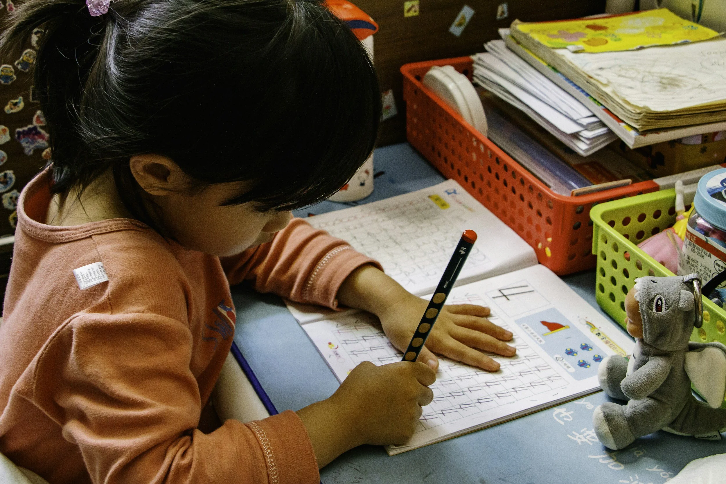 A young girl with dark hair in a pink jacket is practicing calligraphy with a black and colored pen on a worksheet. The desk has colorful storage containers, books, and a plush toy, with stickers on the wall in the background.