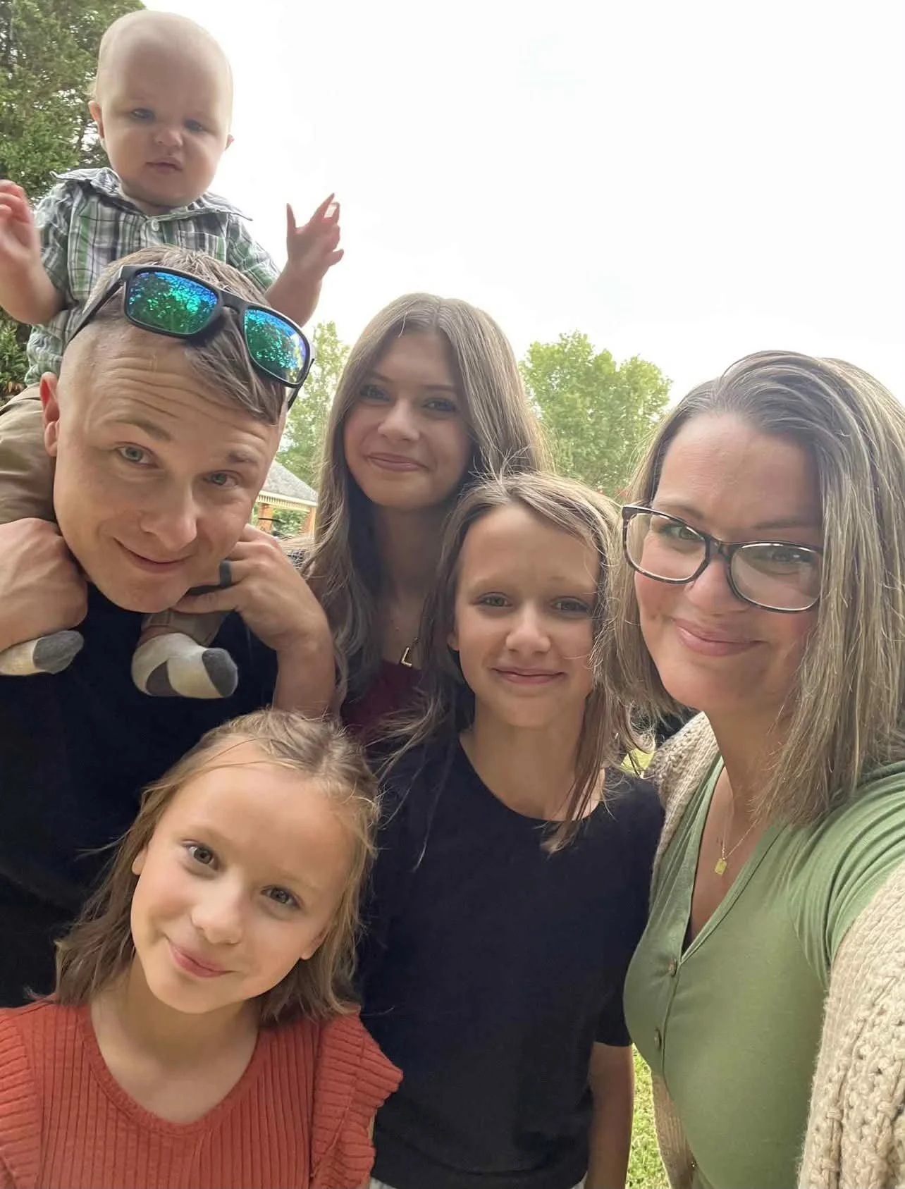 Outdoor family selfie with two adults, three children, and a baby riding on a man's shoulders.