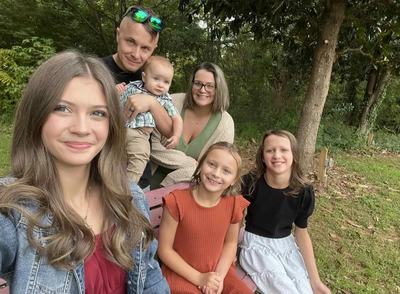 Family of six posing for a selfie outdoors in a wooded area, with a man holding a baby and three girls in front.