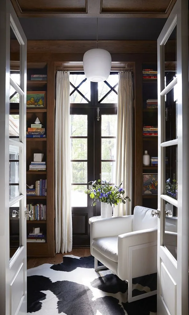 Cozy reading nook with black French doors, white curtains, a white armchair, a black and white cowhide rug, and built-in bookshelves filled with books and decorative items.