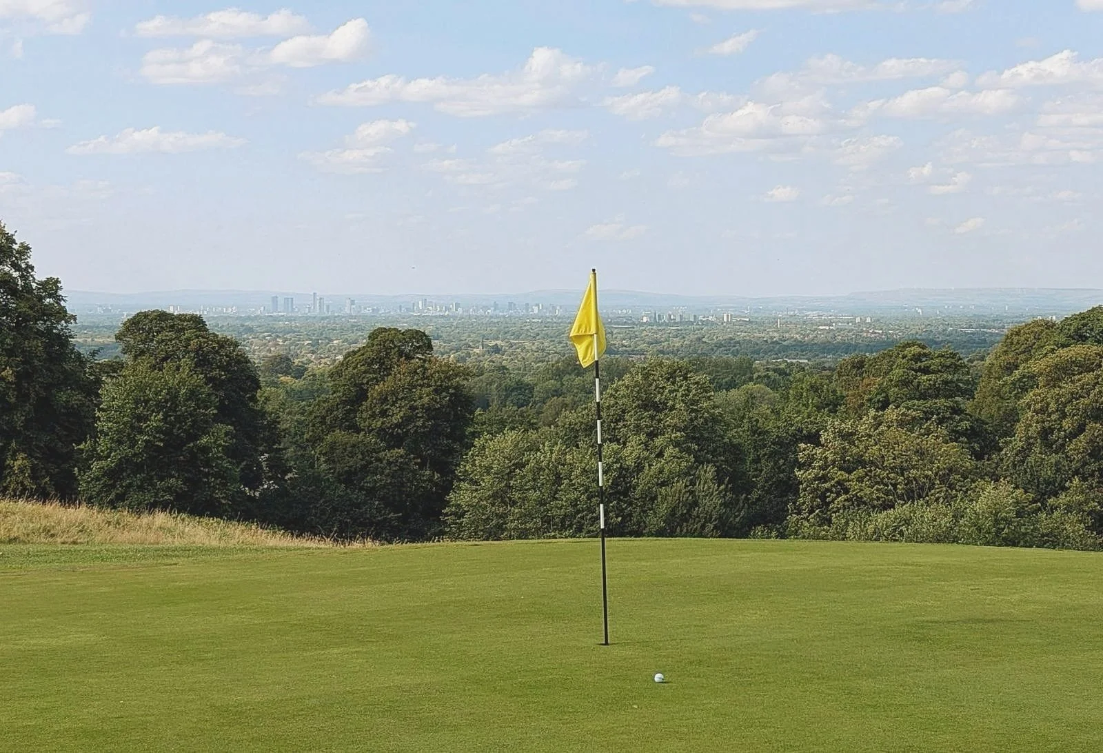 A golf course green with a flagstick and golf ball, surrounded by trees, with a city skyline and hills in the distance under a blue sky with scattered clouds.