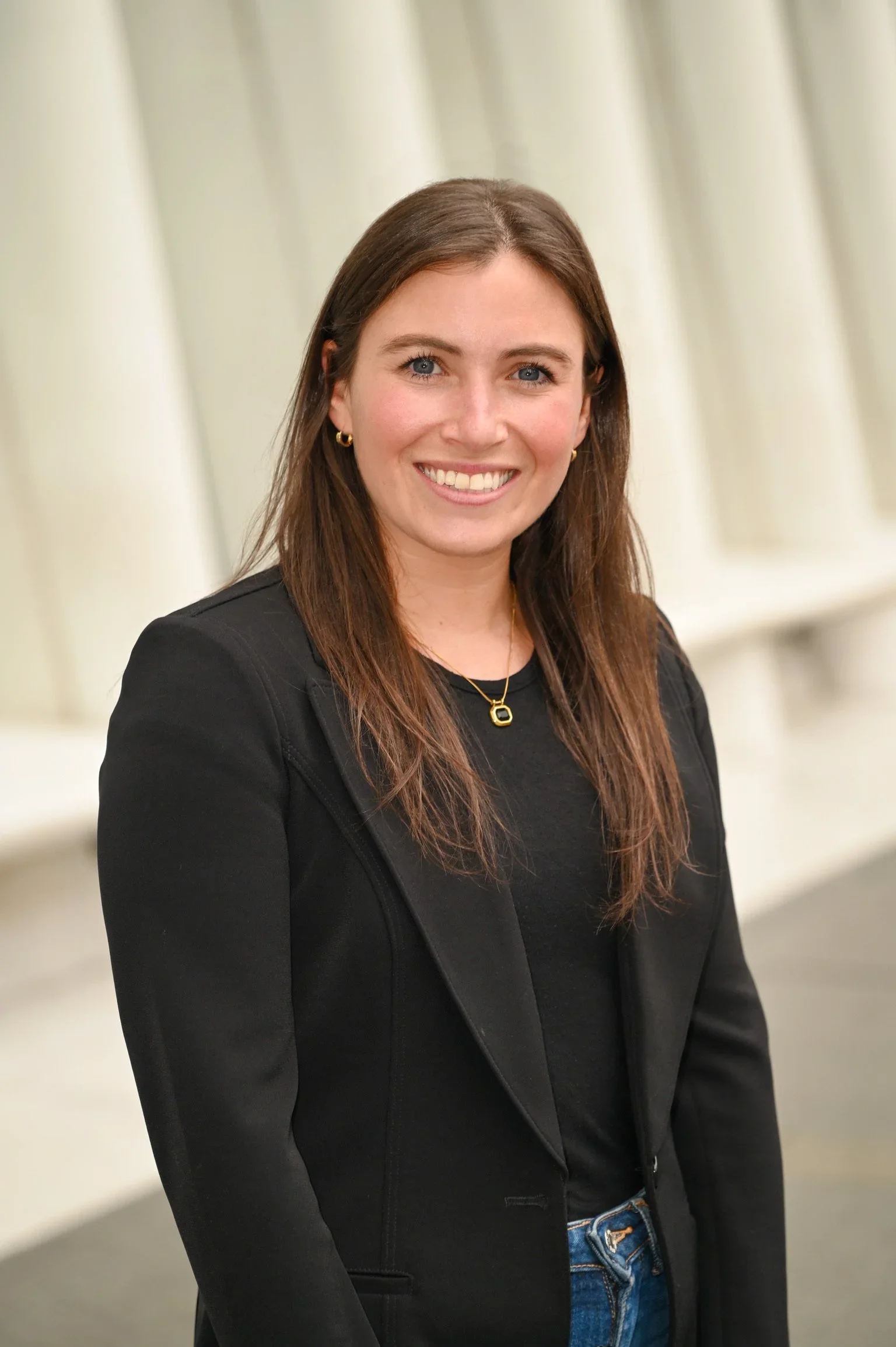 A woman with long brown hair, wearing a black blazer and a black top, standing in front of a cream-colored background, smiling at the camera.