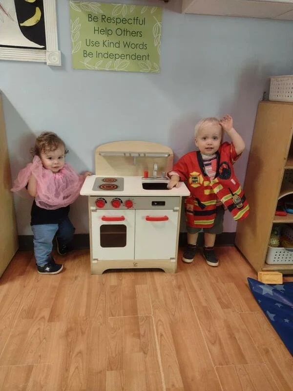 Two young children playing with a toy kitchen. The girl on the left is wearing a pink dress and jeans, and the boy on the right is dressed as a firefighter. They are indoors with a wooden floor, and there is a green sign above them with the words: Be
