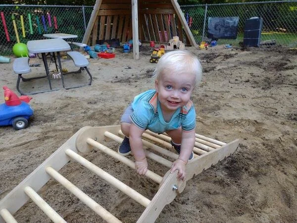 Young child with blonde hair playing on a wooden ladder in a sandbox at a playground.