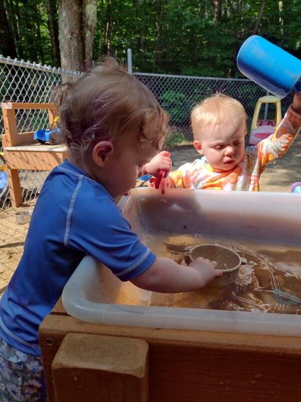Two young children playing at a sand and water table outdoors, with trees and a chain-link fence in the background.