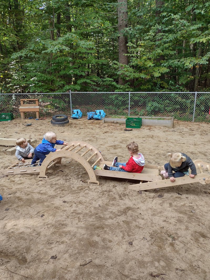 Four children playing on a wooden outdoor playground structure in a sandy area, with a green wooded background and a chain-link fence.