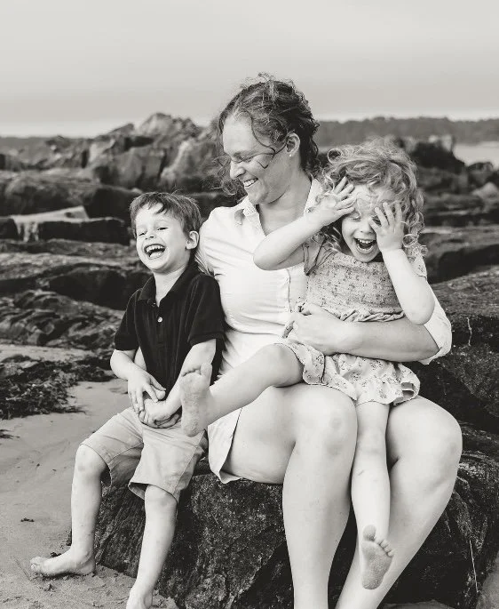 A woman with glasses smiling in front of a plain wall.