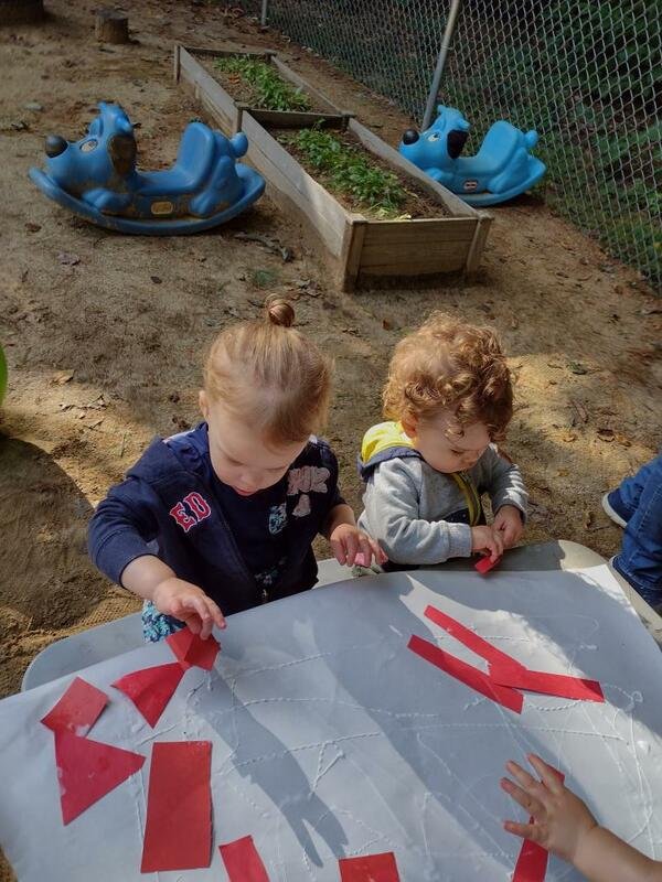 Two young children are sitting at a table outdoors, working with red paper shapes. There are blue rocking toy animals and a garden bed with plants in the background.