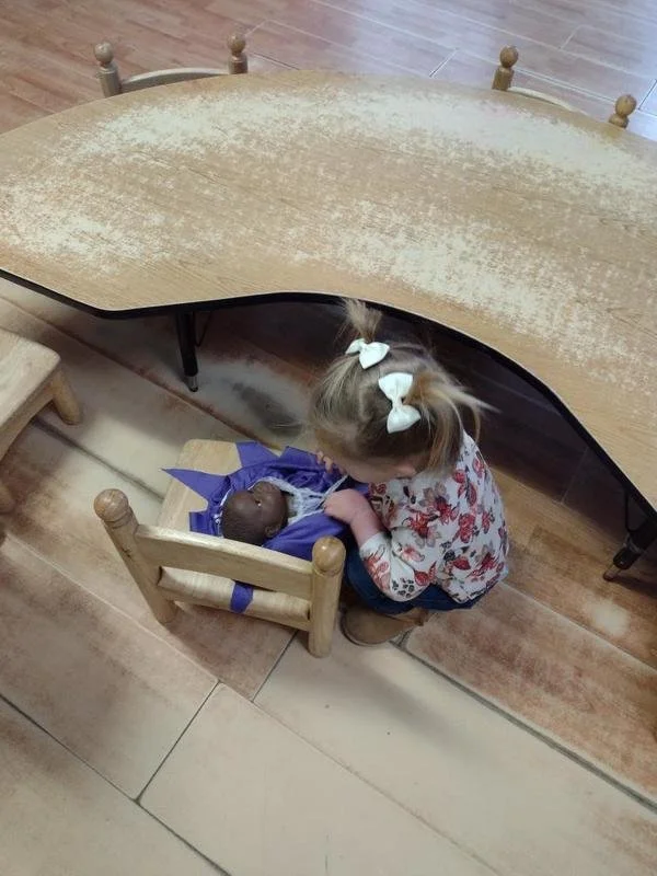 A young girl with bows in her hair kneeling by a small wooden cradle with a doll inside, underneath a large wooden table.