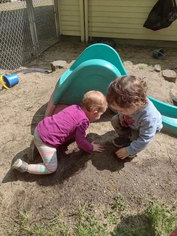 Two young children playing in a sandbox next to a small plastic slide. The girl is wearing a purple long-sleeve shirt and striped leggings, and the boy is wearing a blue hoodie and dark pants. They are both crouched down, digging or drawing in the sa