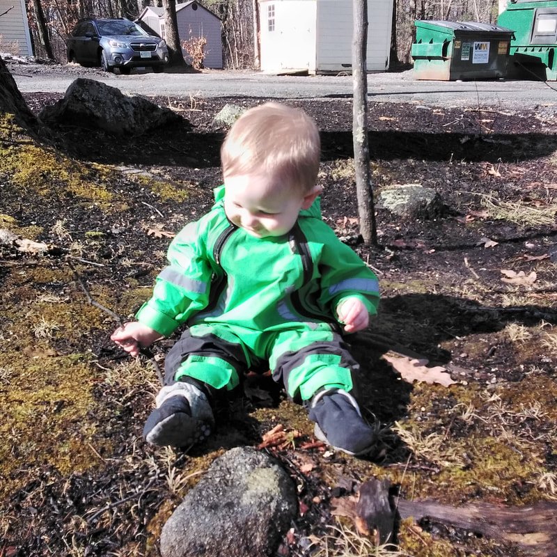 A young child sitting outdoors on the ground, wearing a bright green jacket and black pants, surrounded by dirt and moss, with a tree behind him and cars and green dumpsters in the background.