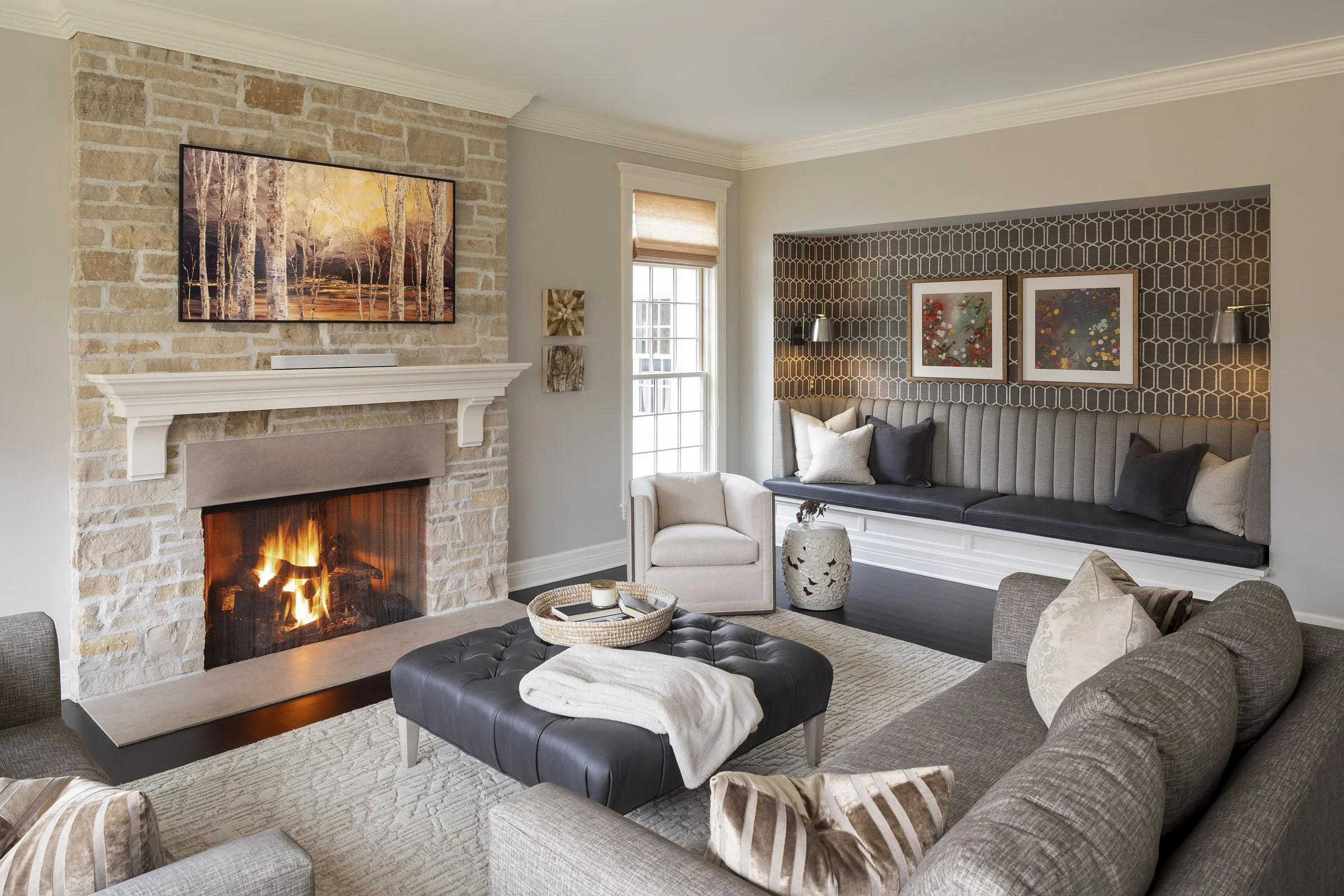Living room with a lit fireplace, gray sofas, a leather ottoman with a tray and blanket, a beige armchair, and a window with a built-in window seat with cushions. There is artwork above the fireplace and on the wall behind the window.