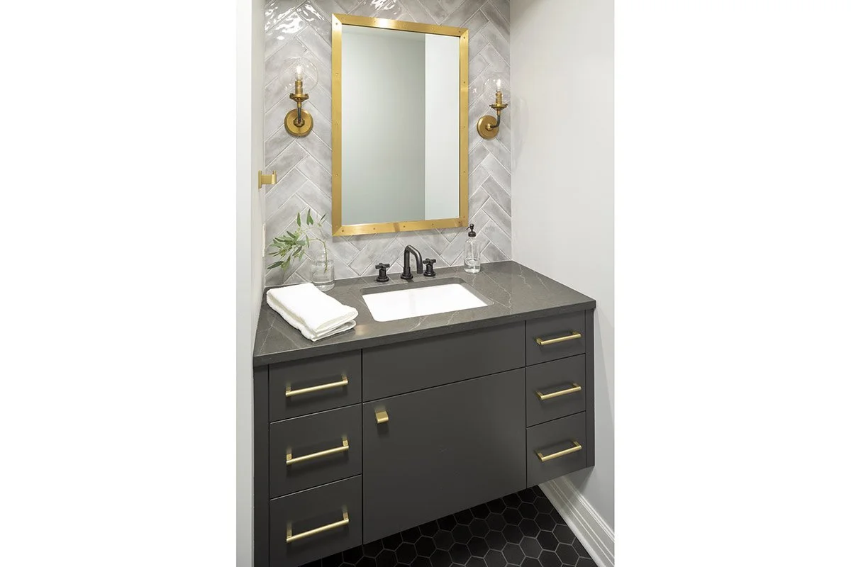 Modern bathroom vanity with black cabinets, gold handles, a gray countertop, a white sink, a mirror framed in gold, and two gold wall sconces.