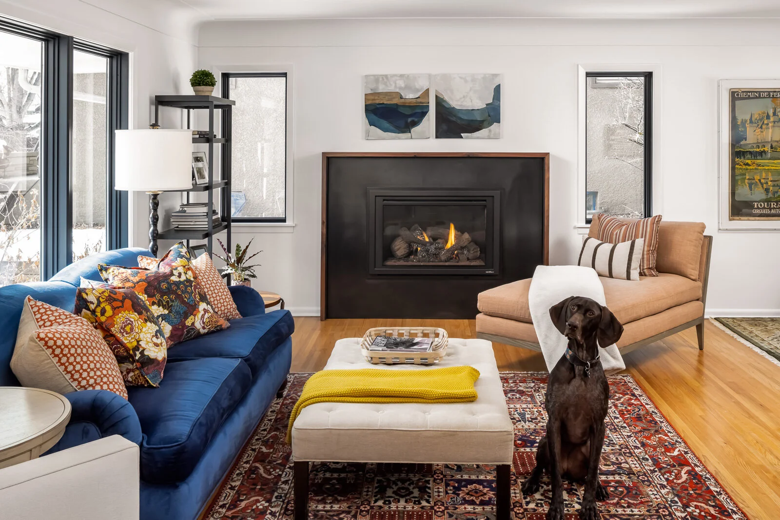 Living room with a blue sofa, a beige chaise lounge, a white ottoman with a yellow throw, a fireplace, and a dog sitting on a Persian rug.