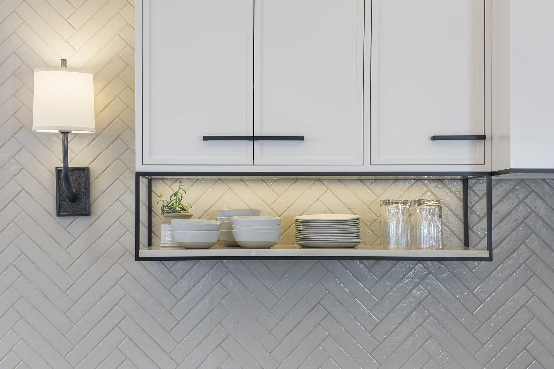 Wall-mounted kitchen shelf with white bowls, plates, and glasses, illuminated by under-cabinet lighting, next to a wall sconce with a white lampshade.