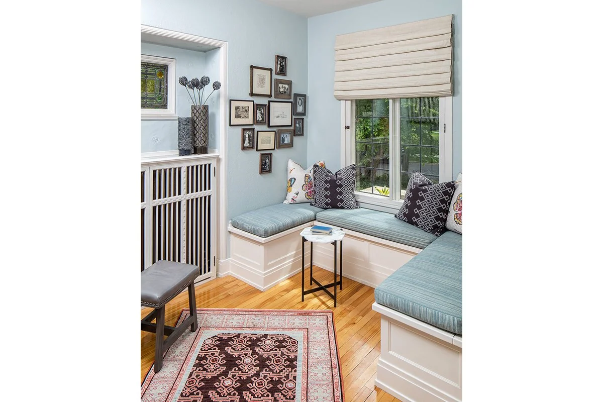 Sunlit window nook with built-in seating, pillows, small side table, framed art on light blue walls, hardwood floor, small window with decorative pane, and patterned area rug.