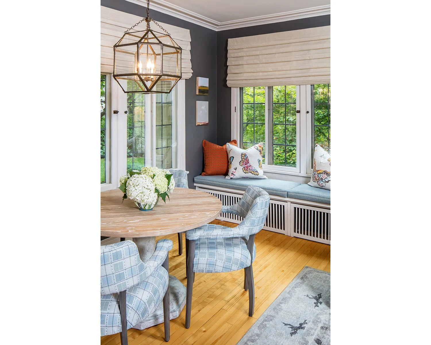 Sunlit dining nook with window seats, cushions with butterfly patterns, a round wooden table with a vase of white flowers, and a geometric pendant light.