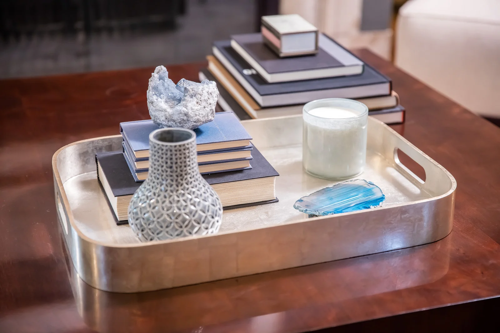 Decorative tray on a wooden table holding stacked books, ceramic vase, candle, and an abstract acrylic art piece.