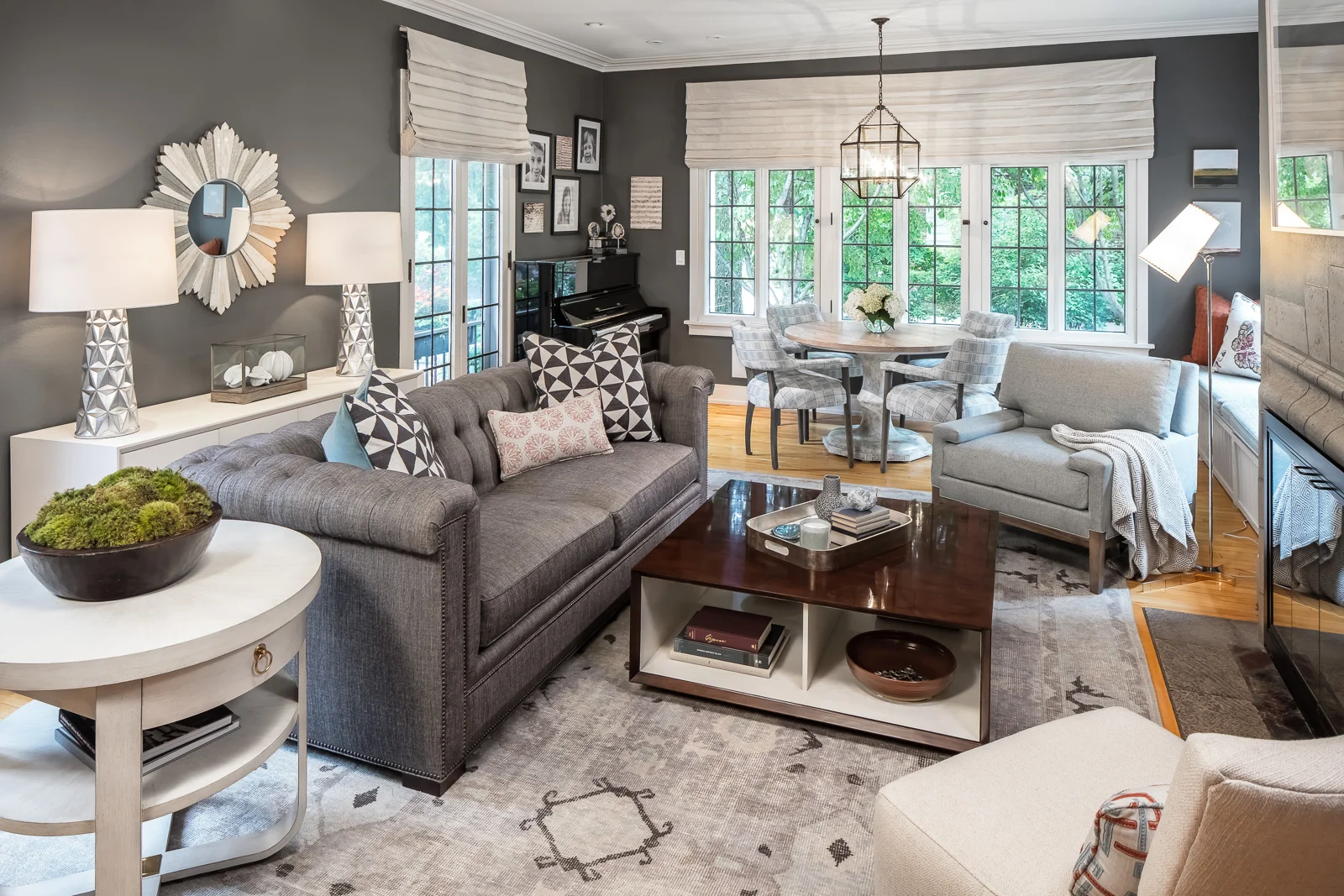 A living room with a gray sofa, a white armchair, a wooden coffee table, and a round dining table with chairs near large windows. The room features gray walls, decorative pillows, lamps, a black upright piano, and various wall art and mirrors.