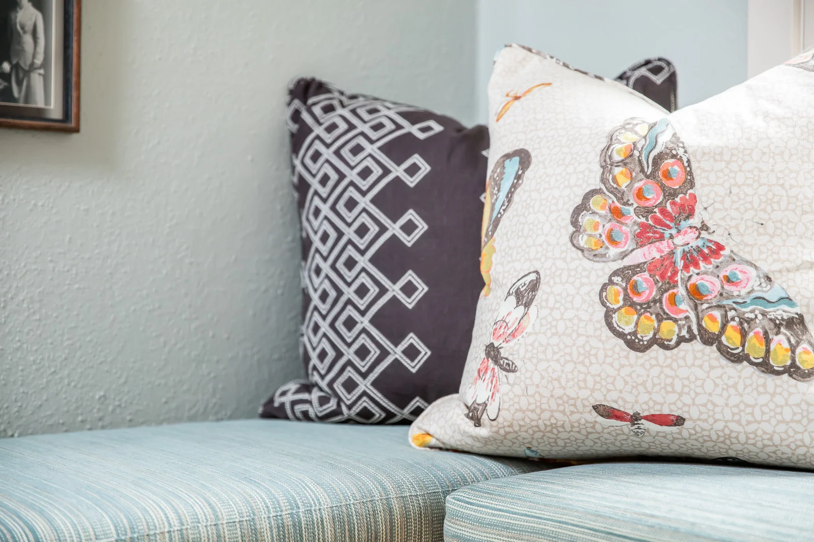 Three decorative throw pillows on a light blue striped bench against a light-colored wall, with a framed black-and-white photograph partially visible on the wall.