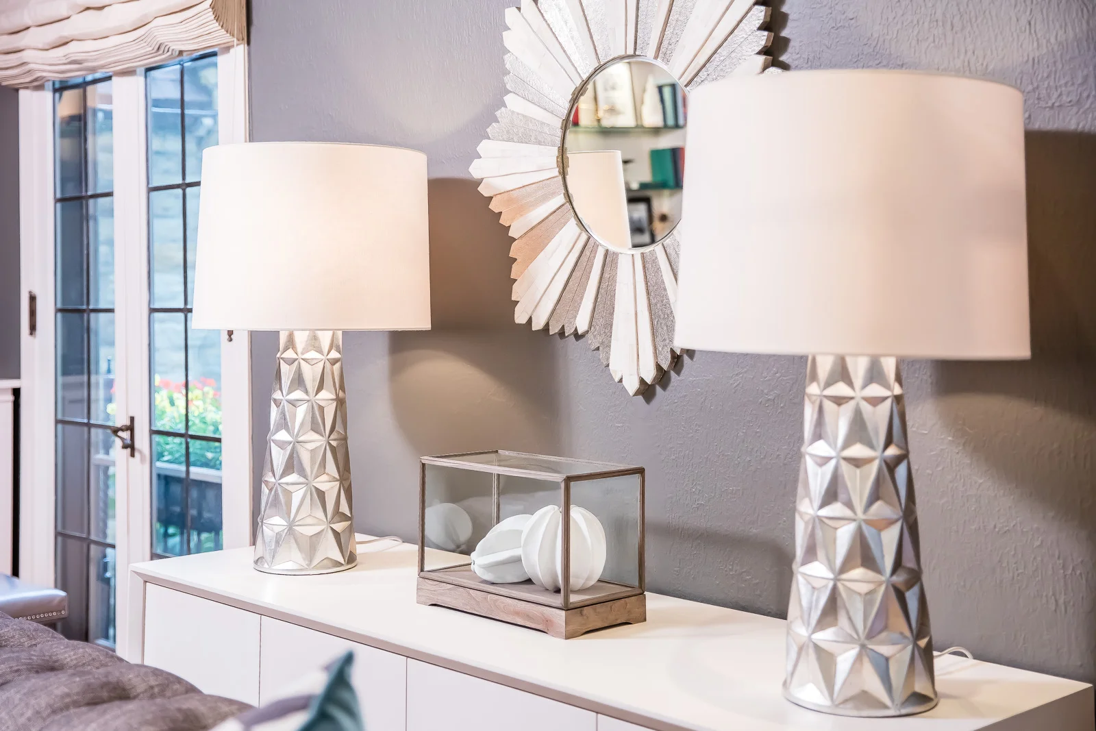Decorative white lamps on a white sideboard with a gray wall, a sunburst mirror, and a glass display box with white ceramic objects. French door with a beige valance in the background.