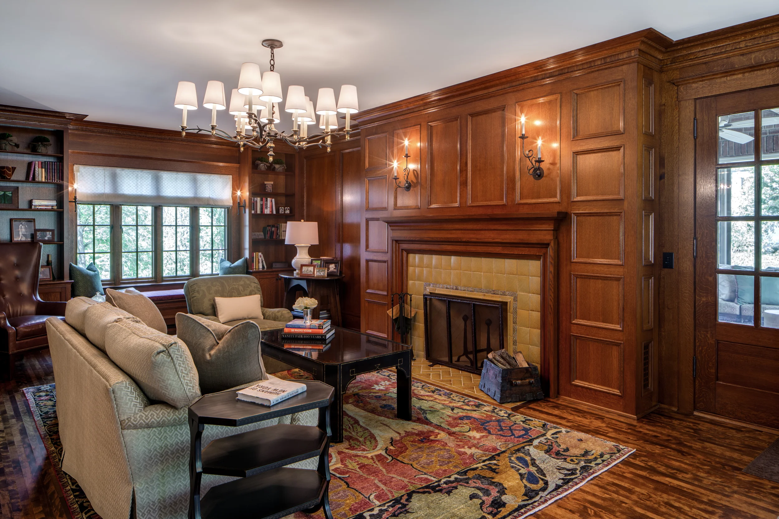Living room with wooden panel walls, a fireplace, a chandelier, and built-in bookshelves, with sofas and a colorful area rug.