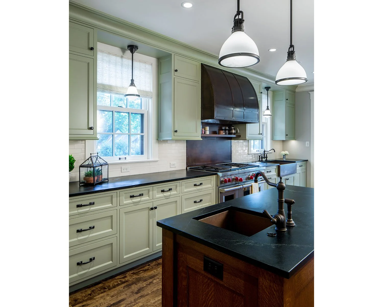 A modern kitchen with light green cabinetry, black countertops, a central island with a dark wood base, and a black stove with a large metal ventilation hood. Windows with white blinds let in natural light, and there are three hanging pendant lights
