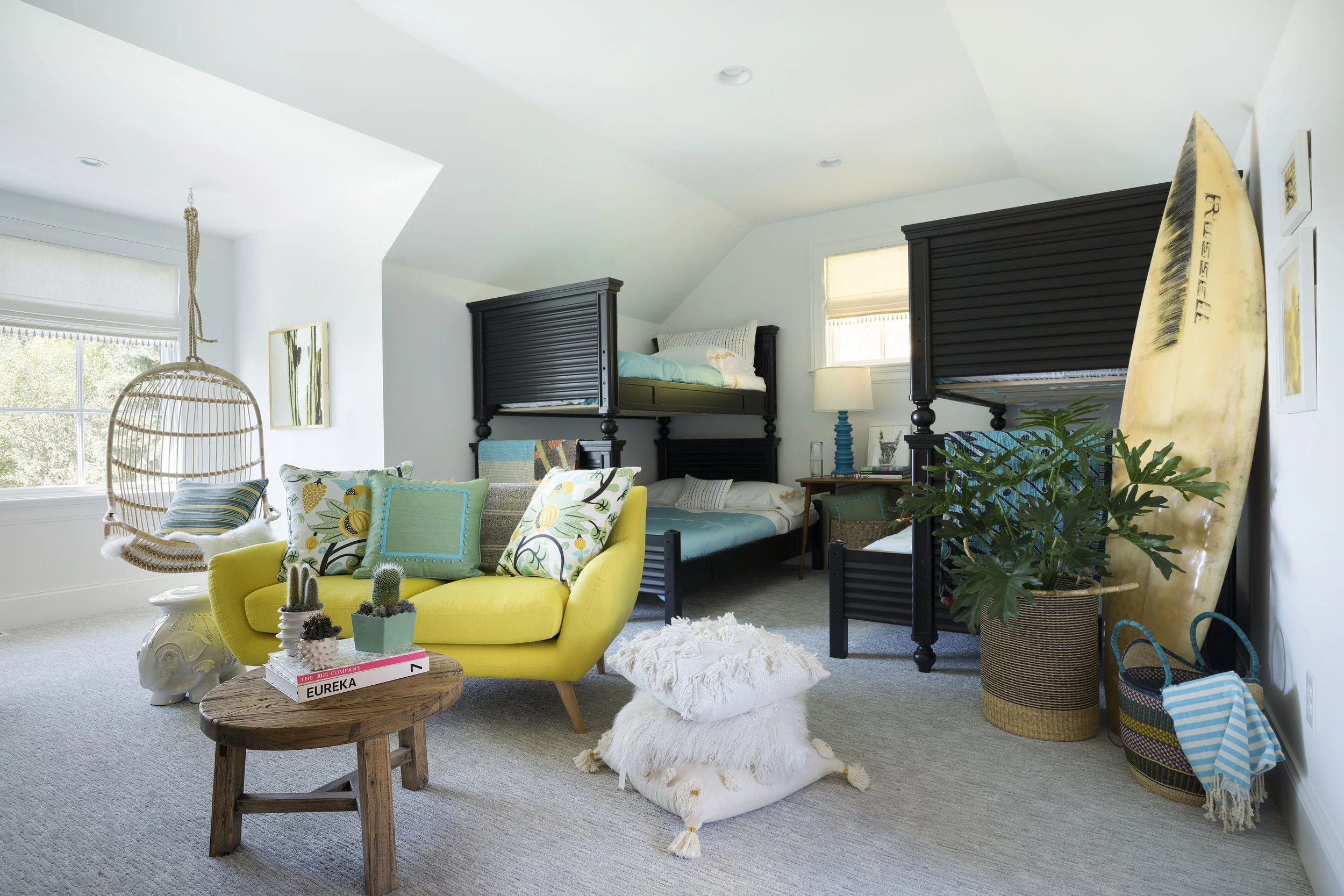A cozy bedroom with bunk beds, a yellow sofa with colorful pillows, a hanging chair, a wooden coffee table with small potted cacti, a large plant in a woven basket, and a surfboard leaning against the wall.