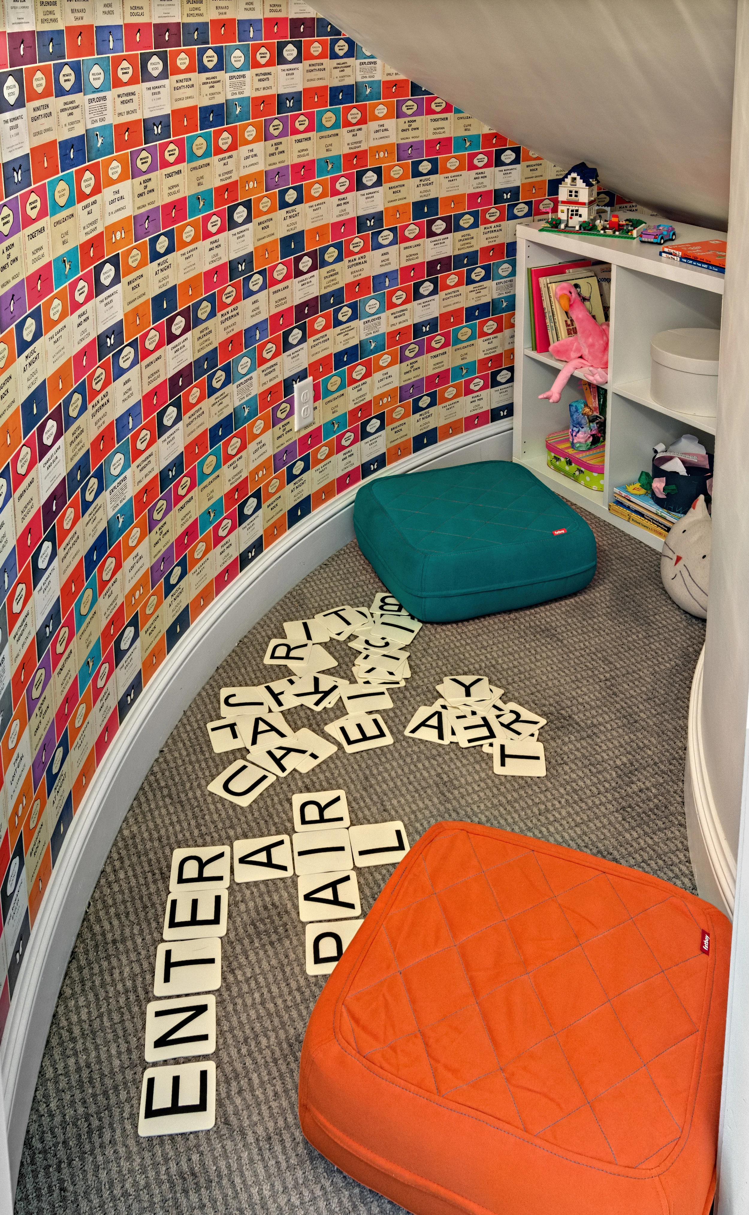 Corner of a child's playroom with colorful wall decorated with alphabet and words, scattered Scrabble tiles on carpet, orange and teal cushions, and white bookshelf holding toys, books, and a pink plush flamingo.