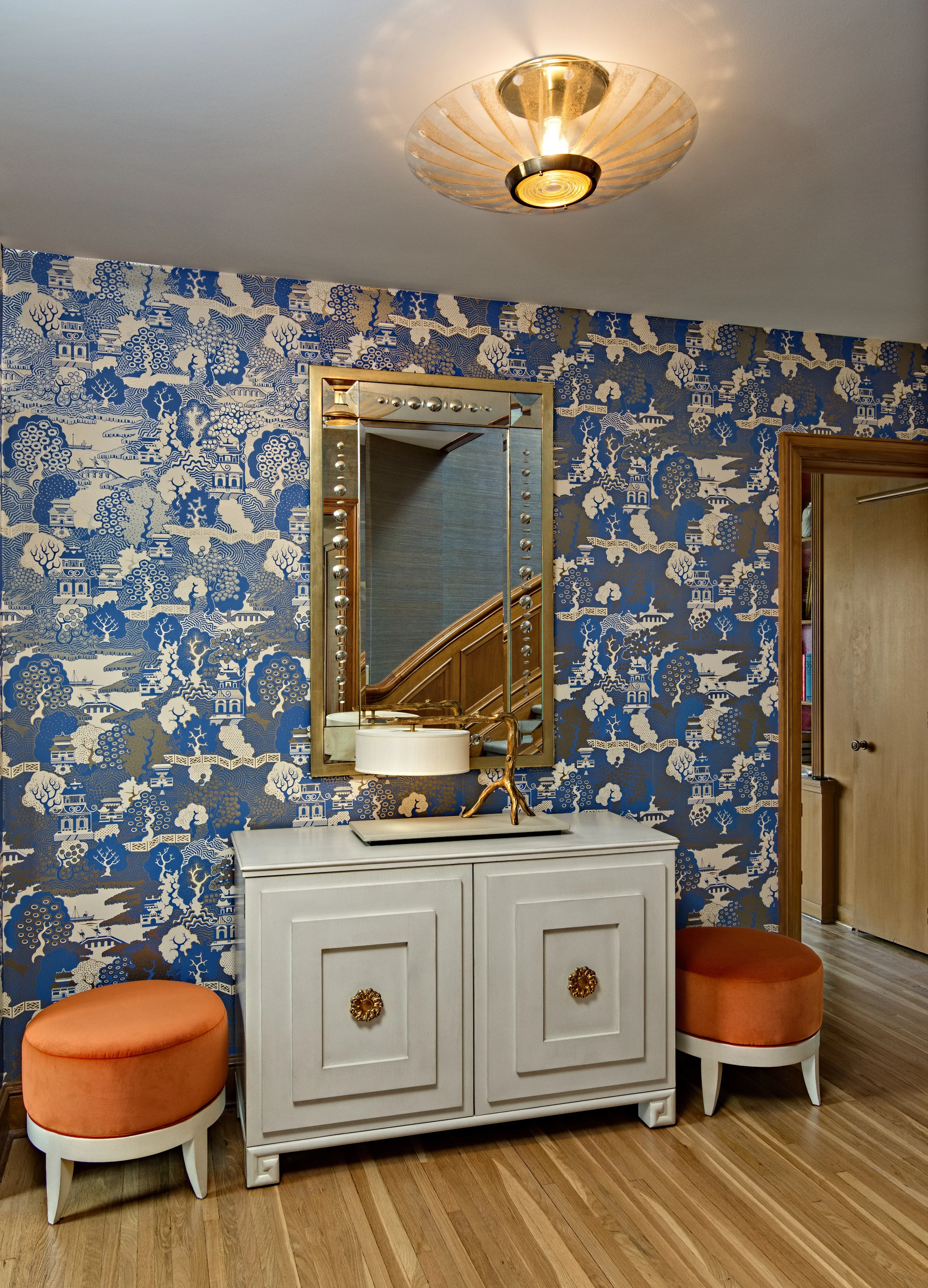 A hallway with blue and white patterned wallpaper, a white cabinet with gold accents, an mirror, a modern table lamp, and two orange cushioned stools. There is a ceiling light fixture.